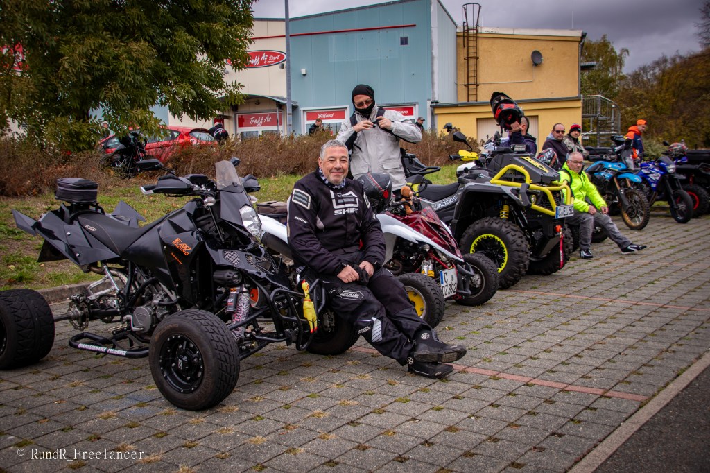 Eine Gruppe von Personen sitzt und steht neben verschiedenen Quads und Motorrädern auf einem Parkplatz. Einer der Fahrer sitzt auf einem schwarzen Quad und lächelt, während andere Fahrer sich in der Nähe aufhalten. Im Hintergrund sind Gebäude und Bäume sichtbar.