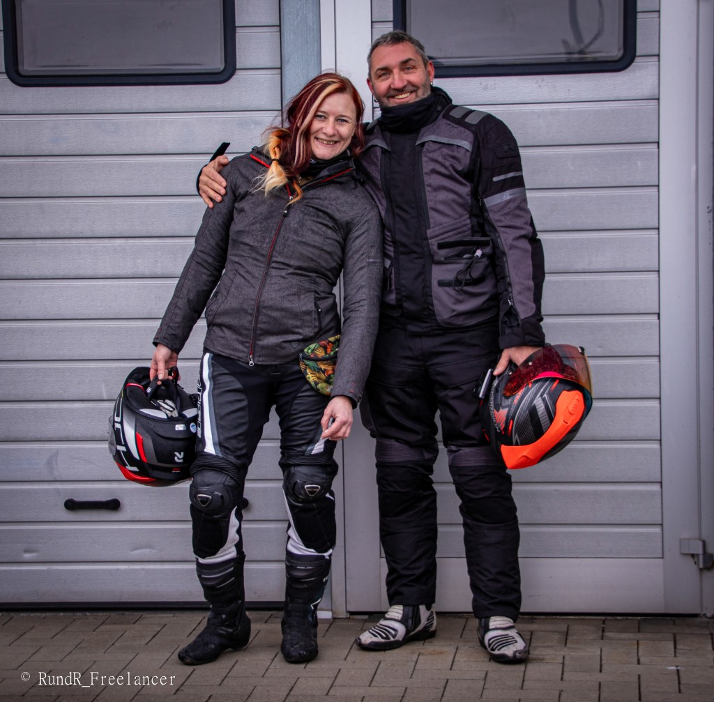 Ein Paar in Motorradkleidung posiert lächelnd vor einer grauen Wand, beide halten Helme in der Hand.