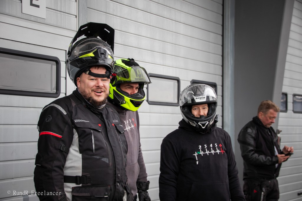 Drei Motorradfahrer in Schutzkleidung und Helmen stehen vor einer Garage. Im Hintergrund ist ein weiterer Mann mit Tätigkeiten beschäftigt.