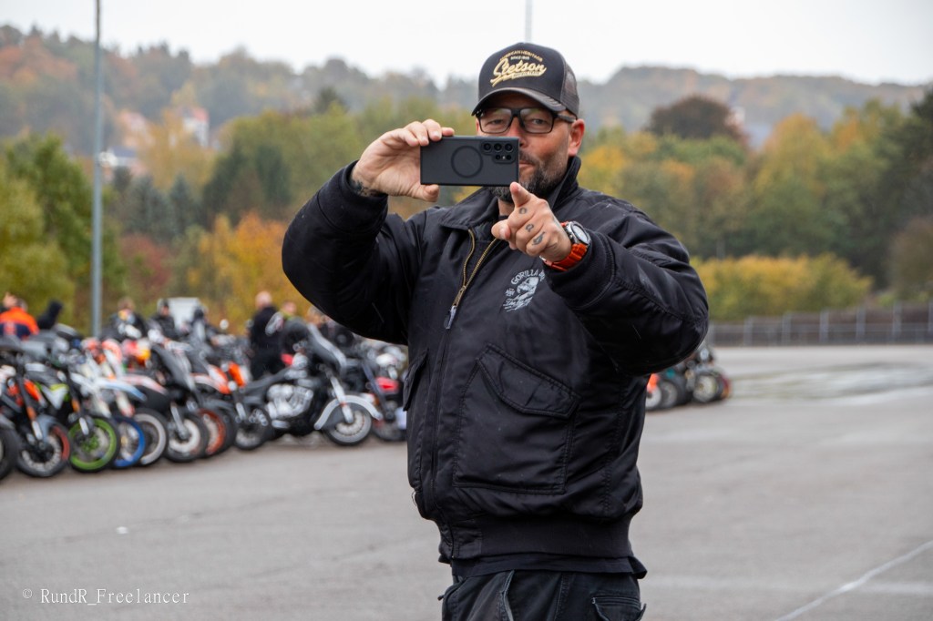 Ein Mann in einer schwarzen Jacke hält ein Smartphone und macht ein Foto, während im Hintergrund Motorräder geparkt sind.