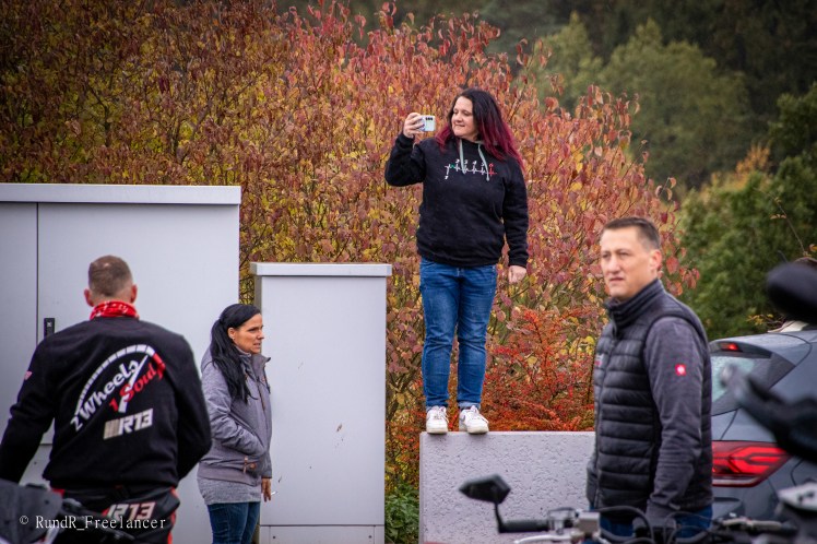 Eine Frau mit bunten Haaren steht auf einer Mauer und macht ein Foto mit ihrem Smartphone, während mehrere Personen in der Nähe stehen und die herbstliche Landschaft im Hintergrund zu sehen ist.