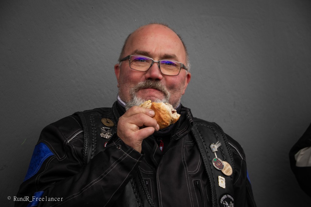 Ein bärtiger Mann in Motorradkleidung hält ein Brötchen und lächelt.