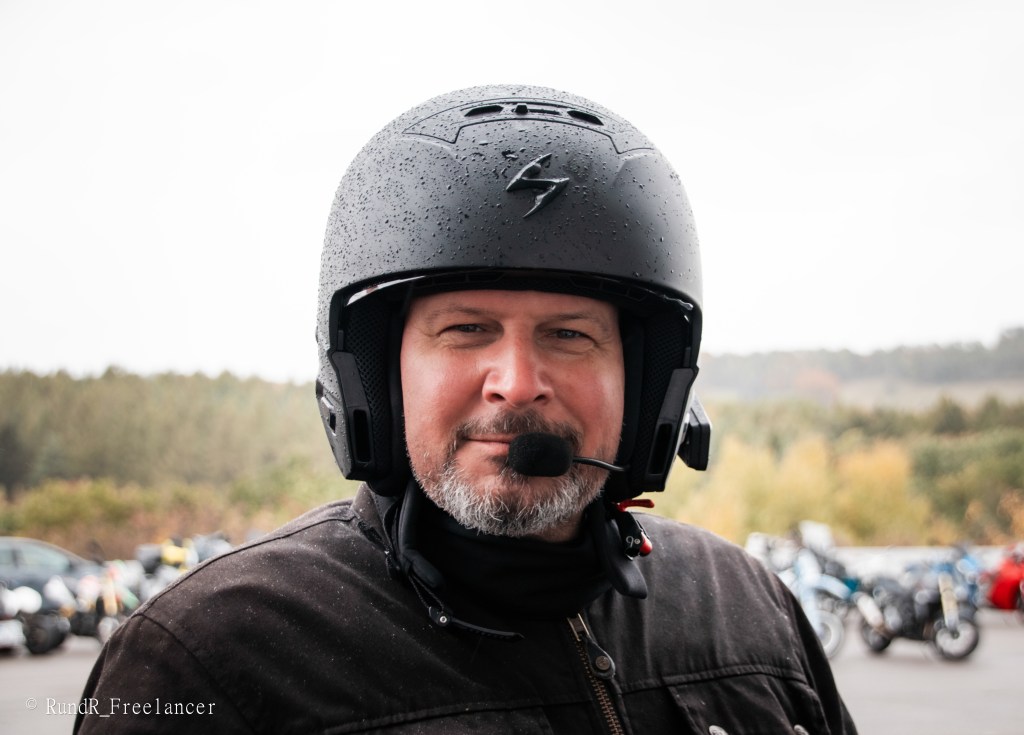 Ein Mann mit einem schwarzen Motorradhelm und einem Mikrofon steht im Freien. Er lächelt in die Kamera, während im Hintergrund Motorräder und eine bewaldete Landschaft zu sehen sind.